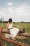 Meisje met blonde haren en zwarte hoed zit op een hek, draagt een lichte jurk en cognac cowboylaarzen.