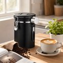 Zwarte RVS koffiebewaarpot op een houten tafel, naast een kopje cappuccino en een zakje koffiebonen, met een plant op de achtergrond.