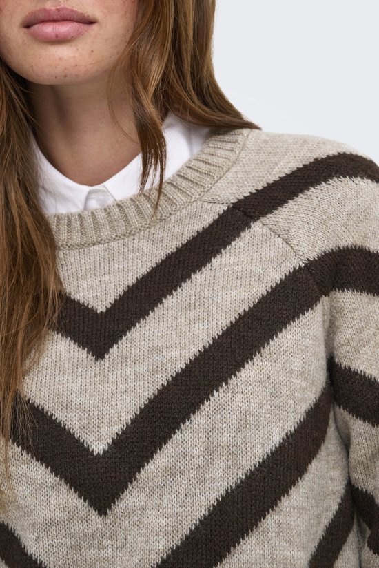 Detailopname van een beige trui met bruine chevronprint, gedragen over een witte blouse.