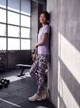 Jong aziatisch model in een lichtpaars sportshirt en een legging met een veelkleurig abstracte print, staand tegen een betonnen muur.