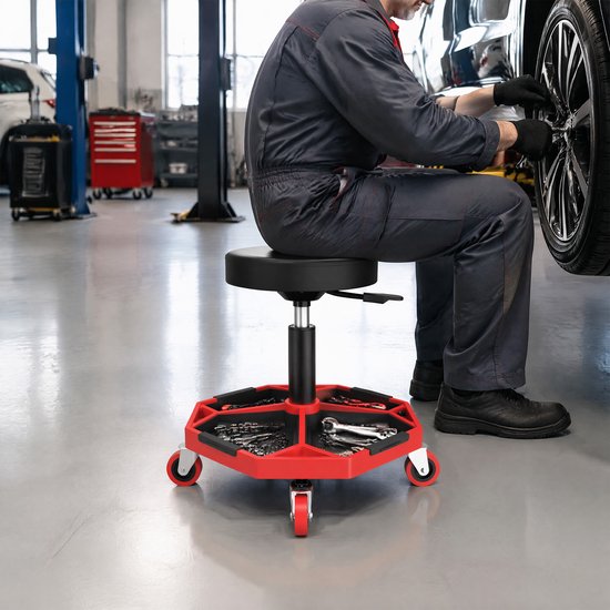 Monteur zittend op een zwarte werkplaatskruk met gereedschapsbak, werkend aan een auto.