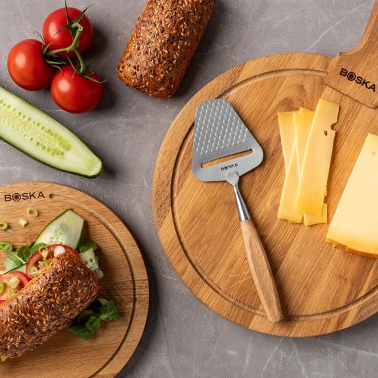 Houten kaasplank met kaasrasp, plakjes kaas en een stuk kaas. Tomaat, komkommer en broodje ernaast.