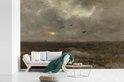 Un salon avec un canapé en bois clair et une table d'appoint, face à un tableau de marais avec un ciel nuageux.