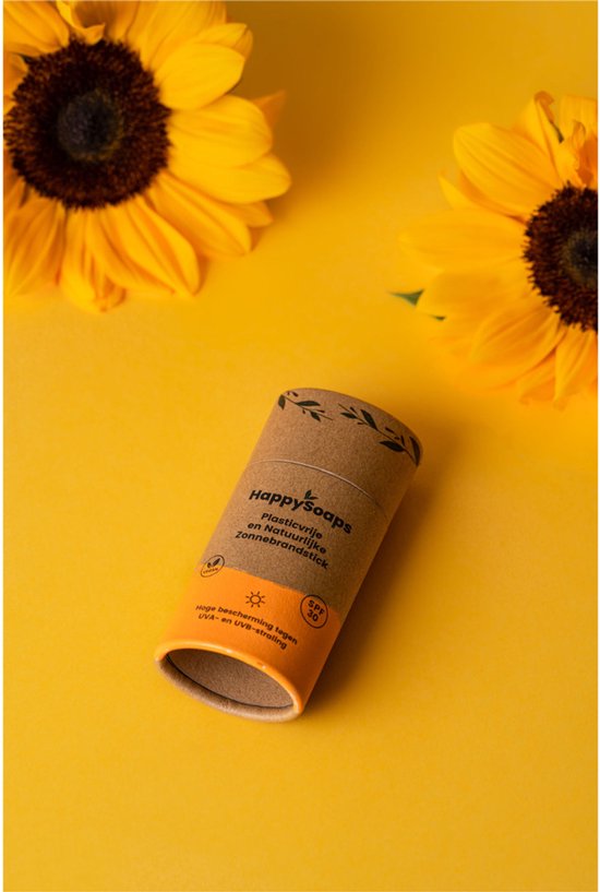 HappySoaps plasticvrije, natuurlijke zonnebrandstick met oranje opdruk, tussen zonnebloemen op gele achtergrond.