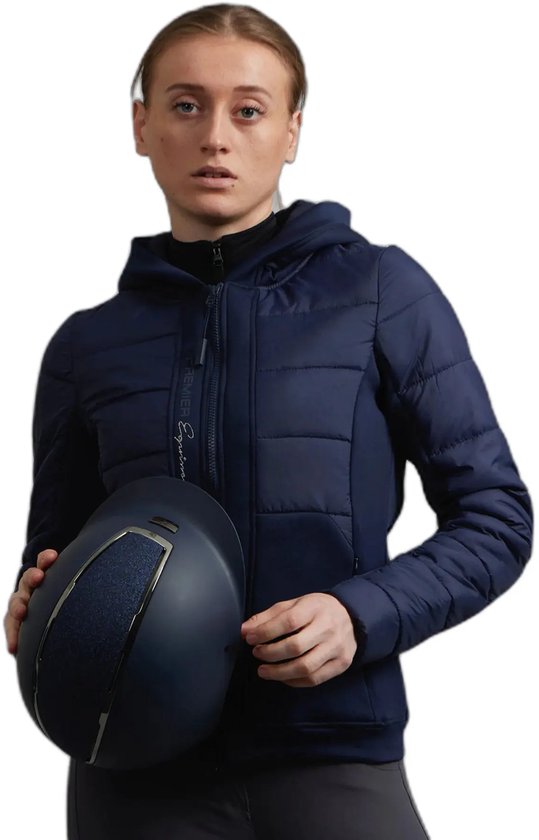 Femme portant une veste d'équitation bleu marine matelassée avec capuche et panneaux latéraux gris. Tenant un casque d'équitation.
