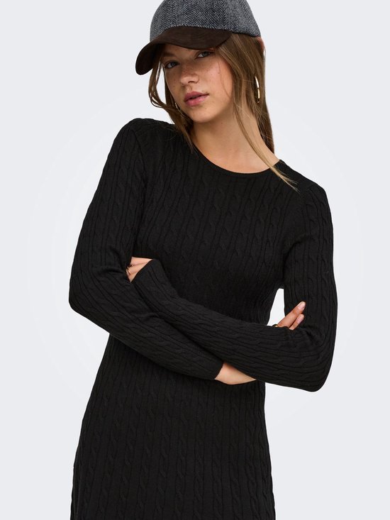 Model met gekruiste armen in een zwarte gebreide mini-jurk met kabels en lange mouwen, met een pet.