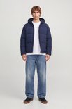 Model draagt een donkerblauwe pufferjack, open rits, met witte T-shirt en jeans.