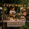Vrouw bij een tafel vol kaarsen en handgemaakte producten, versierd met lampjes en planten.