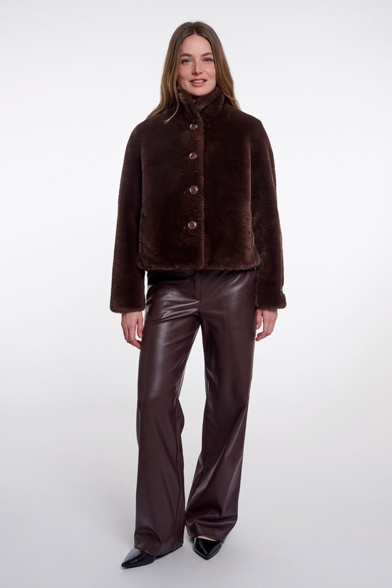 Een model draagt een korte, bruine faux fur jas met een hoge kraag en knopen. De jas is gecombineerd met bruine leren broeken.