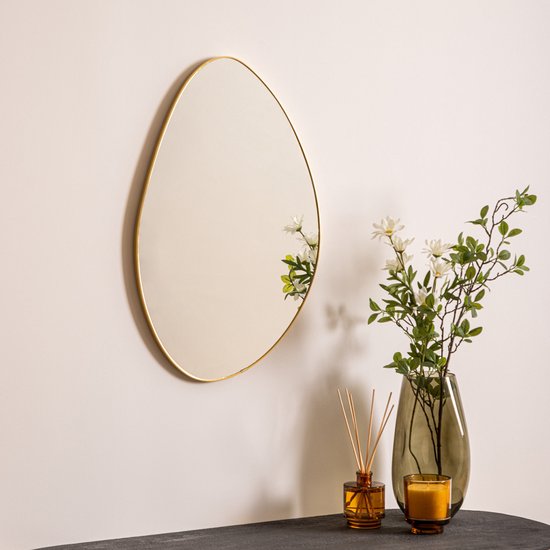 Organische wandspiegel met goudkleurig frame, hangend aan een witte muur naast een vaas met bloemen.