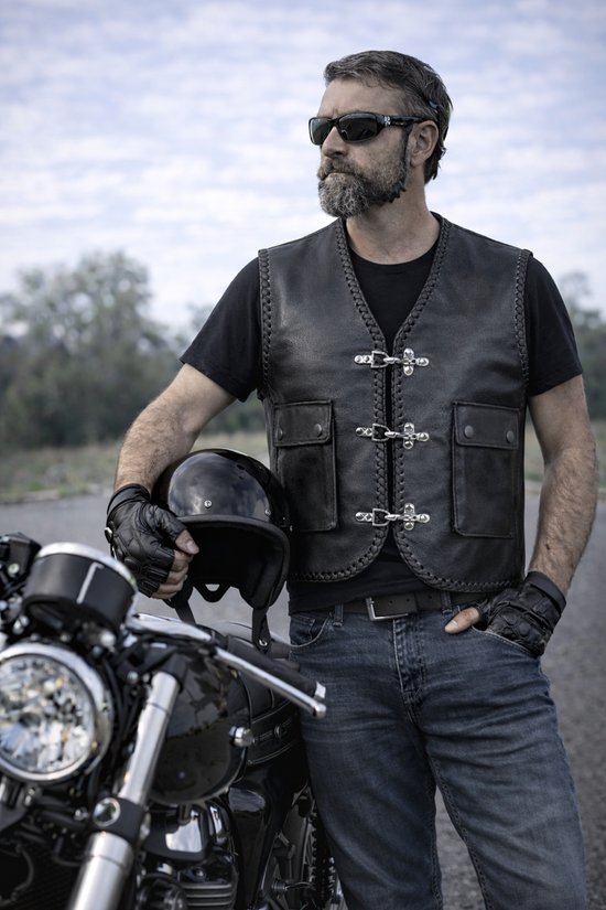 Man met baard en zonnebril draagt een zwart leren bikerjack, helm in de hand, naast een motor.