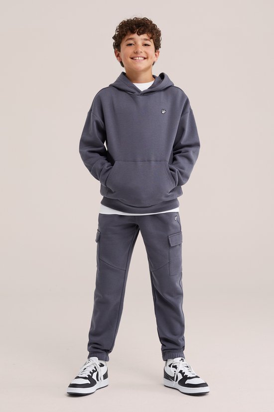 Jongen in antracietgrijze joggingbroek met cargozakken, hoodie en witte t-shirt. Sneakers met zwart-witte details.