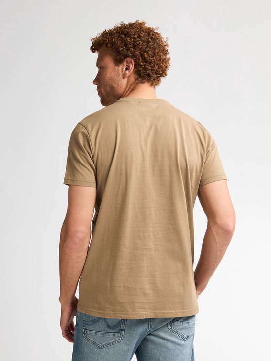Man met krullend rood haar, gedraaid naar links, draagt lichtbruin t-shirt en spijkerbroek.