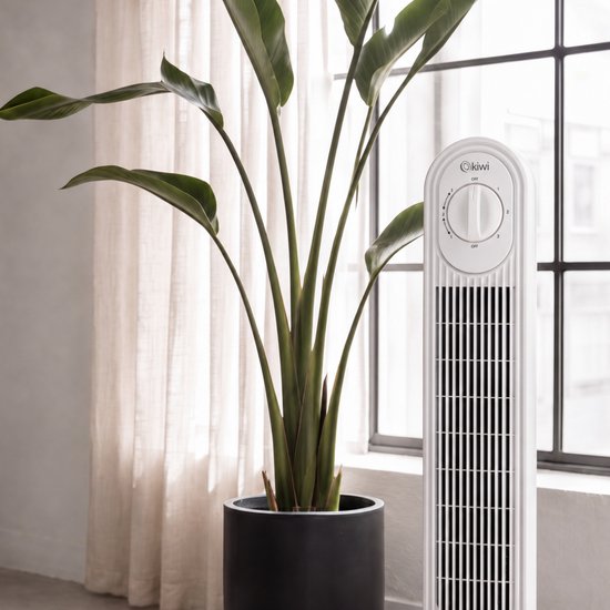 Witte torenventilator naast een grote groene kamerplant en achtergordijnen.