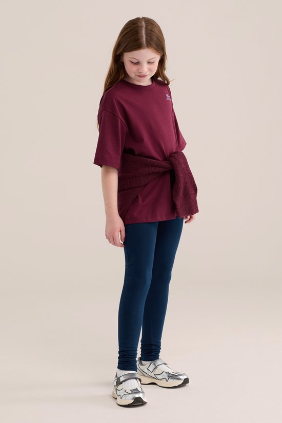 Meisje in donkerblauwe legging en bordeauxrood t-shirt met trui om de heupen.