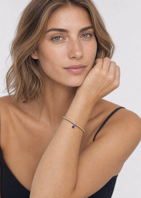 Model met een zwarte top, draagt een gouden armband met blauwe hanger.