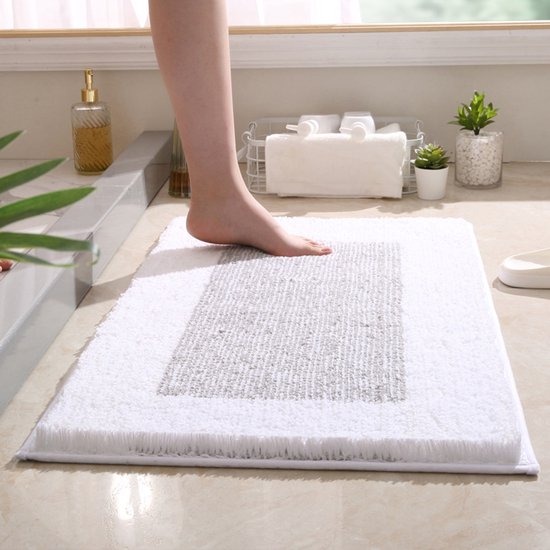 Voet op een witte badmat met grijze textuur in een badkamer met planten en zeepdispenser.