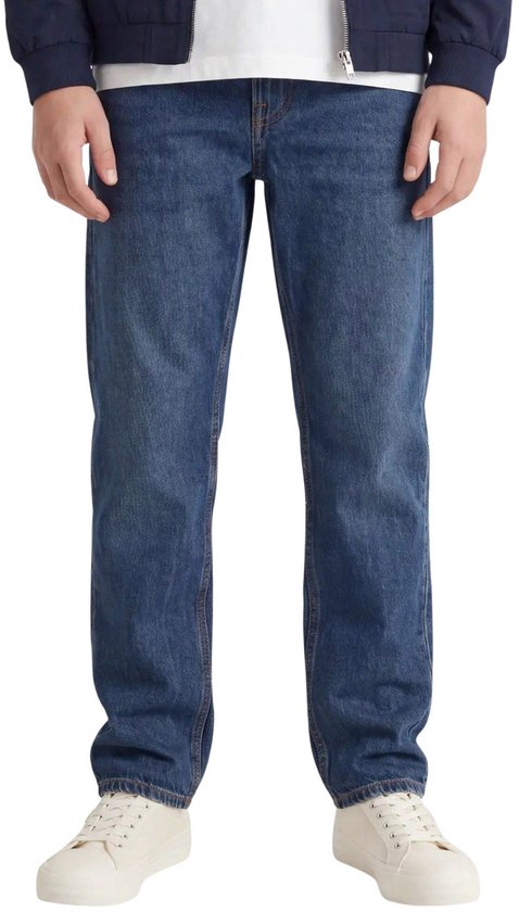 Onderlichaam van een jongen in een wit T-shirt, blauwe jeans en witte sneakers.