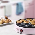 Roze poffertjespan met een rode aan/uitknop. Er liggen poffertjes op de pan en een stapel poffertjes op een schaal ernaast.