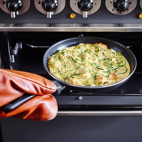 Een oranjebruine ovenwant pakt een pan met courgetteschotel uit een oven.