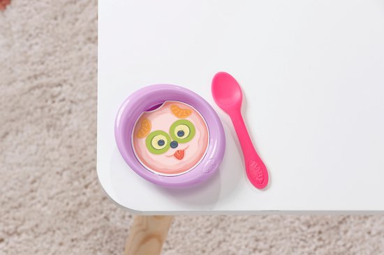 Paars bakje met babyvoeding met gezicht van kiwi en sinaasappel, naast een roze lepel.