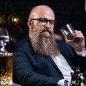 Man met lange baard en bril, gekleed in geruit pak, zit in een leren stoel en houdt een glas whiskey vast.
