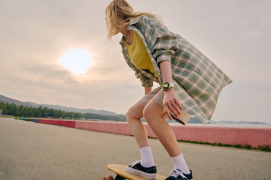 Een meisje met een geel shirt en groen geruit overhemd, rijdend op een longboard bij zonsopgang.