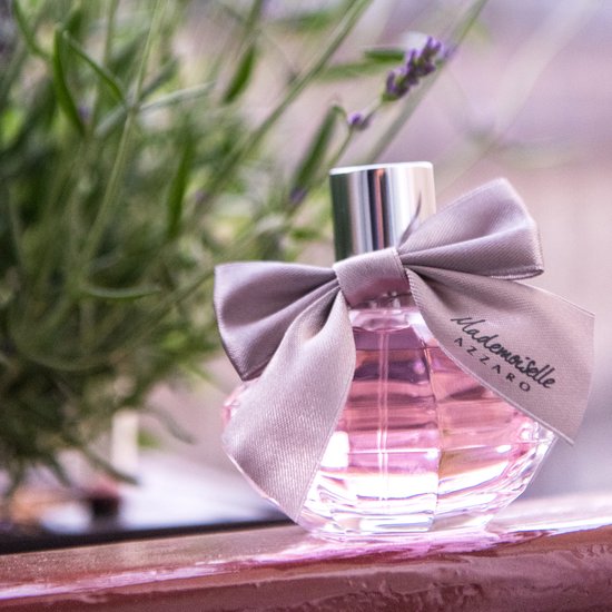 Flasque de parfum Mademoiselle Azzaro avec liquide rose et nœud argenté, avec de la lavande en arrière-plan.