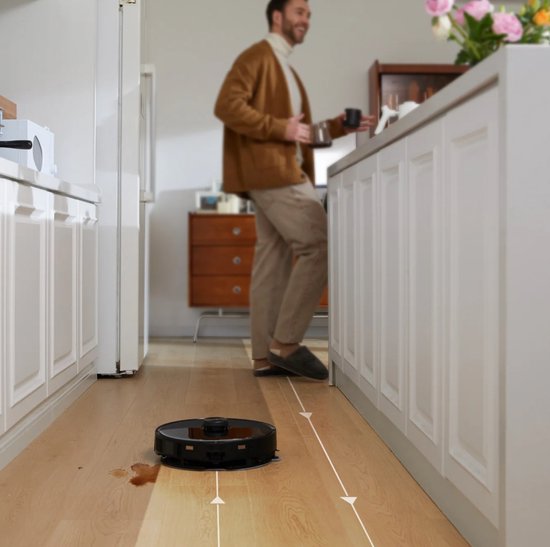 Zwarte robotstofzuiger zuigt vlekken op een houten vloer in een keuken, man staat op achtergrond.