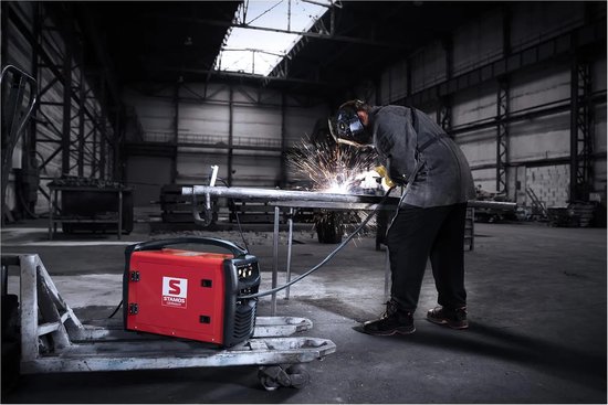 Persoon last een metalen pijp in een grote werkplaats, met een rood lasapparaat op de voorgrond.