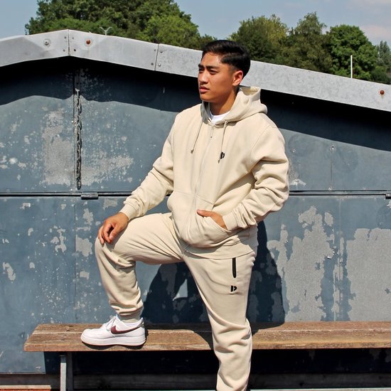 Man poseert in een zandkleurige joggingbroek en hoodie, zittend op een bankje.