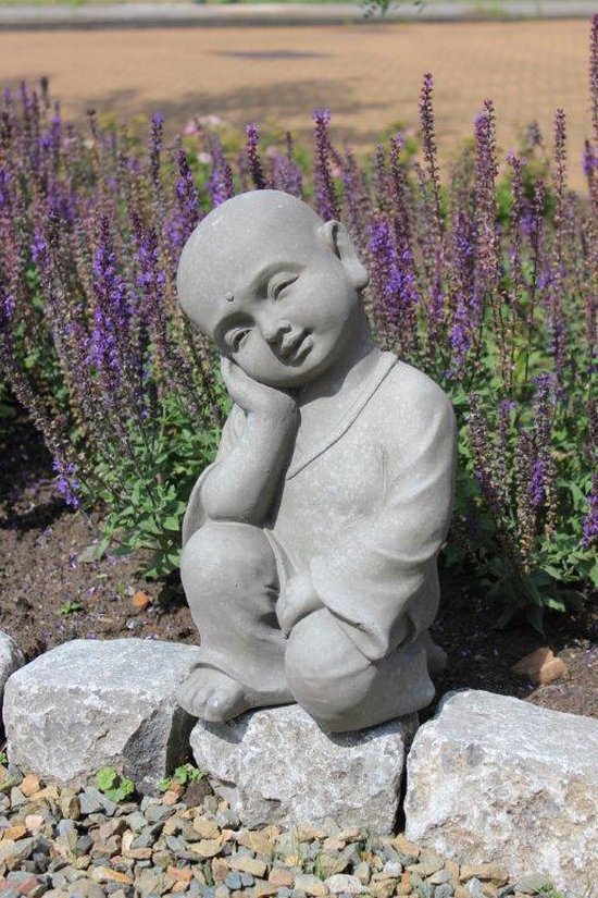 Statue de Bouddha enfant en pierre grise, assis en tailleur, tête sur la main, entre des fleurs violettes.