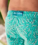 Close-up van de achterkant van een turquoise zwembroek met wit patroon en merklabel 'SEA'SONS'.
