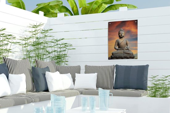 Bouddha en pierre sur fleur de lotus sur fond de coucher de soleil orange, suspendu à une clôture blanche.