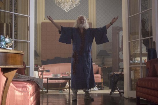 Een man met een baard en lang grijs haar staat in een kamer met zijn armen geheven, gekleed in een blauw gewaad.
