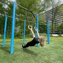 Vrouw met blonde haren in zwart sportkleding oefent aan KRAKEN gymringen aan blauwe palen.
