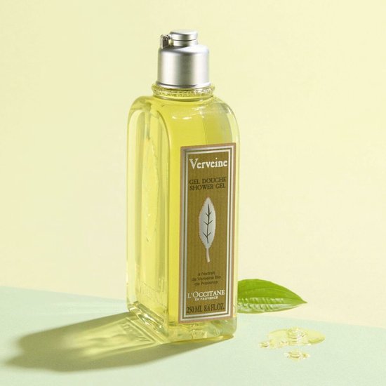 Vierkante, doorzichtige fles met gele douchegel, een zilveren dop, en een groen en beige label met een bladprint. Naast de fles ligt een groen blad en een paar druppels gel.