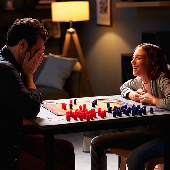 Een vader en dochter spelen Stratego en lachen, met het speelbord en de rode en blauwe pionnen op tafel.
