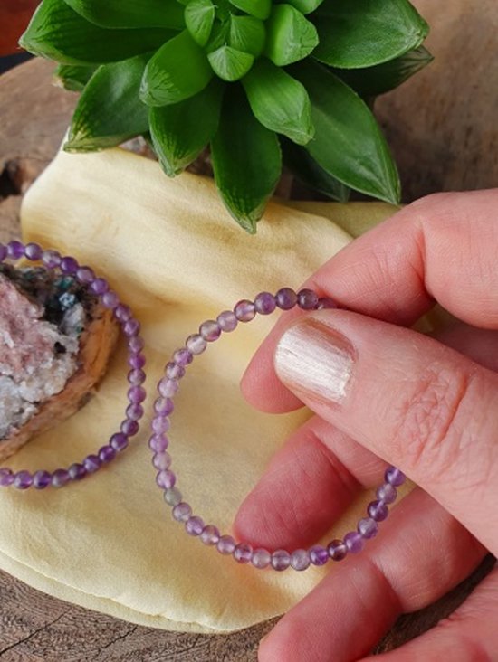 Hand houdt een armband vast van paarse amethistkralen, met een rots en plant op de achtergrond.