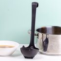 Zwarte soeplepel in de vorm van een katje met pootjes, naast een pan soep en een kookpot.