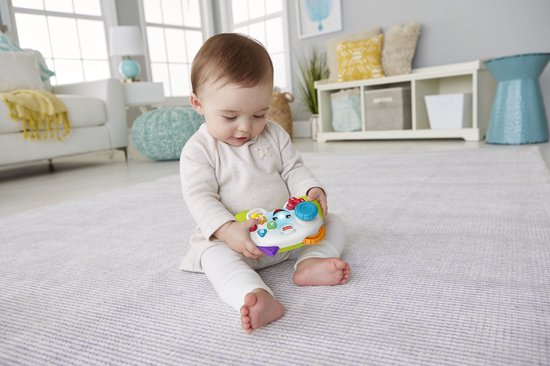 Baby met krullen zit op vloerkleed met Fisher-Price spelcontroller in handen. Speelgoed is groen, wit en heeft gekleurde knoppen.