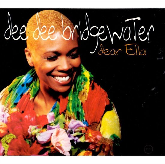 Dee Dee Bridgewater, met kort blond haar, lachend, met een kleurrijke sjaal en een boeket bloemen.