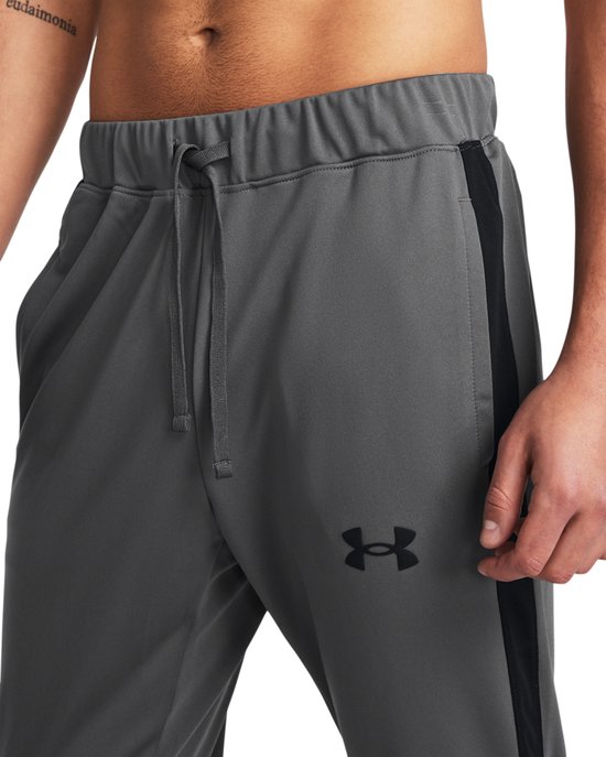 Grijze trainingsbroek met trekkoord, zwarte streep op de zijkant en een zwart Under Armour logo.