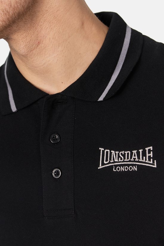 Close-up van een zwarte Lonsdale polo met geborduurd logo 'Lonsdale London' en dubbele grijze strepen op de kraag.