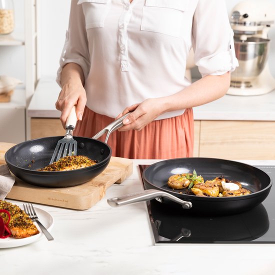 Persoon gebruikt een spatel in een zwarte KitchenAid-pan met vis, naast een pan met gebakken aardappels.