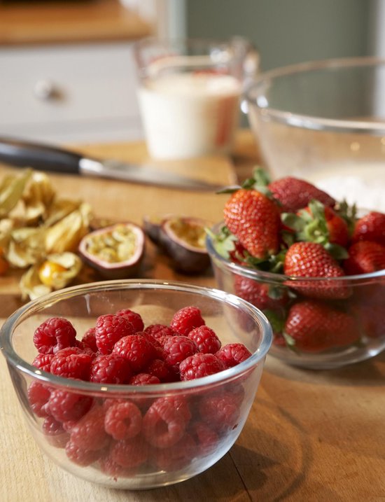 Glazen kom gevuld met frambozen, met een kom met aardbeien en ander fruit op de achtergrond.