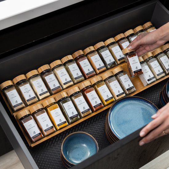 Een keukenlade vol georganiseerde kruidenpotjes op houten rekjes. Een hand pakt een potje. Blauwe borden en bowls liggen aan de zijkant.