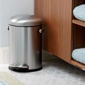 RVS pedaalemmer met ronde deksel en handvatten, staand op een grijze tegelvloer naast een houten kast met blauwe handdoeken.