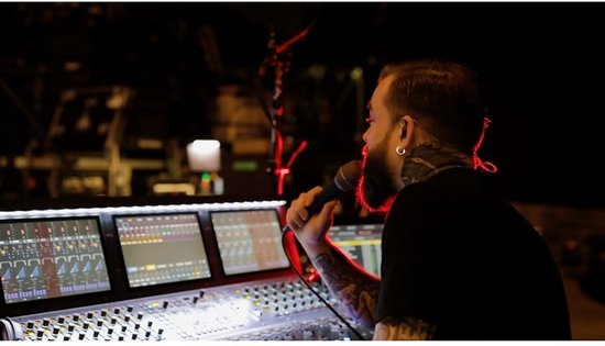 Man met baard en tatoeages zingt in zwarte microfoon met rode grill bij een mengpaneel.