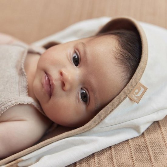 Bébé aux yeux marron, drapé dans une cape de bain blanc cassé douce avec une bordure beige. Le logo Jollein est visible.